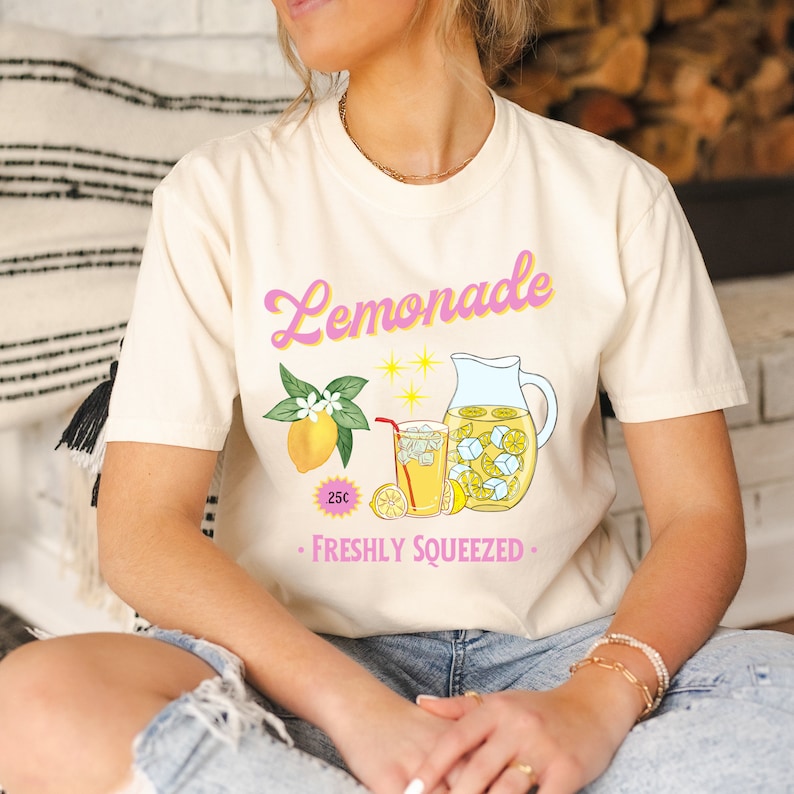 May include: A white t-shirt with a pink "Lemonade" graphic. The graphic features a pitcher and glass of lemonade with lemons, ice cubes, and a red straw. The text "25c" and "Freshly Squeezed" are also included in the graphic.