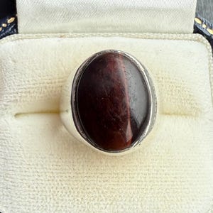 May include: A silver ring featuring an oval, reddish-brown gemstone, presented in a blue jewellery box with a gold clasp. The box has a white interior and a gold-coloured border.