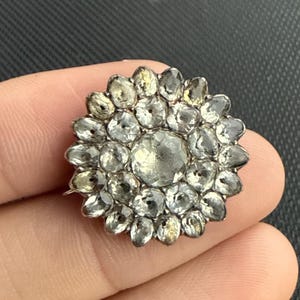 May include: A silver-toned, flower-shaped brooch with a central, large, clear stone surrounded by smaller, clear stones. The brooch has a detailed, vintage design, and the stones appear to be faceted.