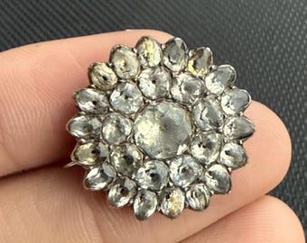 May include: A silver-toned, flower-shaped brooch with a central, large, clear stone surrounded by smaller, clear stones. The brooch has a detailed, vintage design, and the stones appear to be faceted.