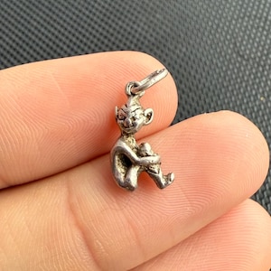 Puede incluir: Un pequeño dije plateado con la forma de un elfo o duende sentado. El dije tiene detalles finos, incluyendo orejas puntiagudas y una expresión caprichosa. Está suspendido de un pequeño lazo, listo para ser añadido a un collar o pulsera.