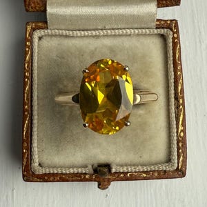 May include: A gold ring featuring a large, oval-cut yellow gemstone, presented in a vintage brown jewellery box with a cream interior. The ring has a thin gold band. The box is open, showcasing the ring in the centre.