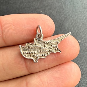 Op de afbeelding: Een kleine, zilverkleurige bedel in de vorm van het eiland Cyprus. De bedel heeft een lus aan de bovenkant om aan een ketting of armband te bevestigen. De namen van steden zijn op de bedel gegraveerd.