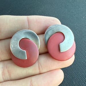 May include: A pair of silver and red earrings. Each earring features a silver, semi-circular piece overlapping a red, circular base. The earrings have a modern, geometric design.