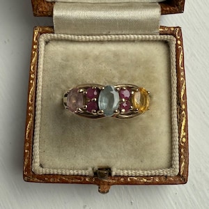 May include: A gold ring set with a line of colourful gemstones, including pink, red, blue, and yellow stones. The ring is presented in an open, vintage brown jewellery box with a beige interior. The box is open, displaying the ring.