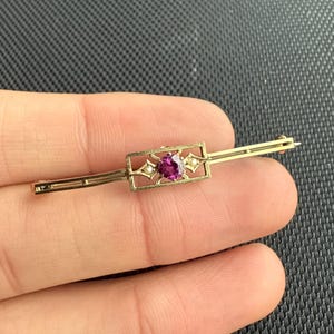 Puede incluir: Un broche de barra dorado con un centro rectangular que presenta una vibrante gema magenta de corte ovalado. El centro está flanqueado por pequeños detalles blancos, similares a perlas. Una pieza de joyería clásica para añadir elegancia.