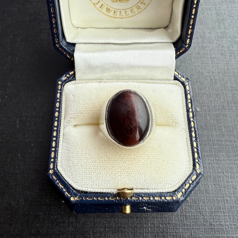 May include: A silver ring featuring an oval, reddish-brown gemstone, presented in a blue jewellery box with a gold clasp. The box has a white interior and a gold-coloured border.