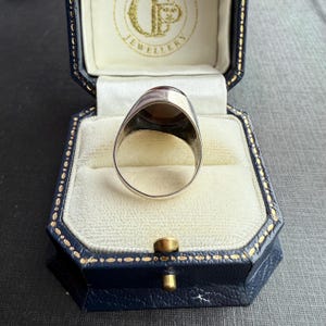 May include: A silver signet ring featuring a large, oval, amber-coloured stone. The ring is presented in a blue jewellery box with a white interior. The box has gold trim and a gold clasp. The ring is a signet style.