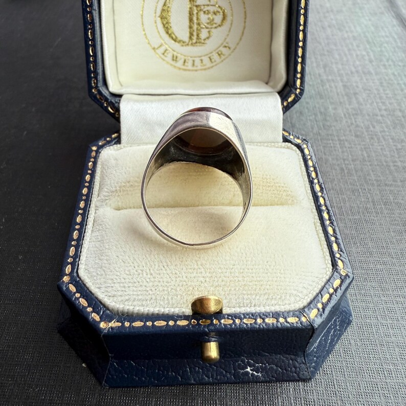 May include: A silver signet ring featuring a large, oval, amber-coloured stone. The ring is presented in a blue jewellery box with a white interior. The box has gold trim and a gold clasp. The ring is a signet style.