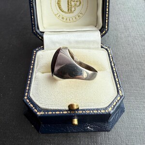 May include: A silver signet ring with a dark red rectangular stone, presented in a blue jewellery box with gold detailing. The ring has a polished surface. The box is open, showing a white interior.