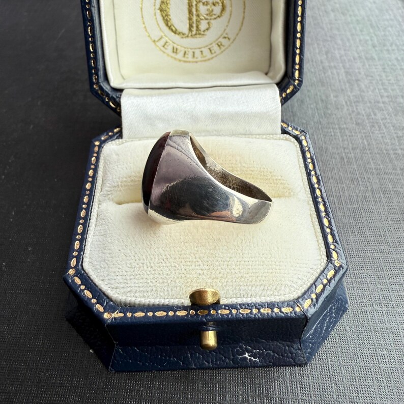 May include: A silver signet ring with a dark red rectangular stone, presented in a blue jewellery box with gold detailing. The ring has a polished surface. The box is open, showing a white interior.