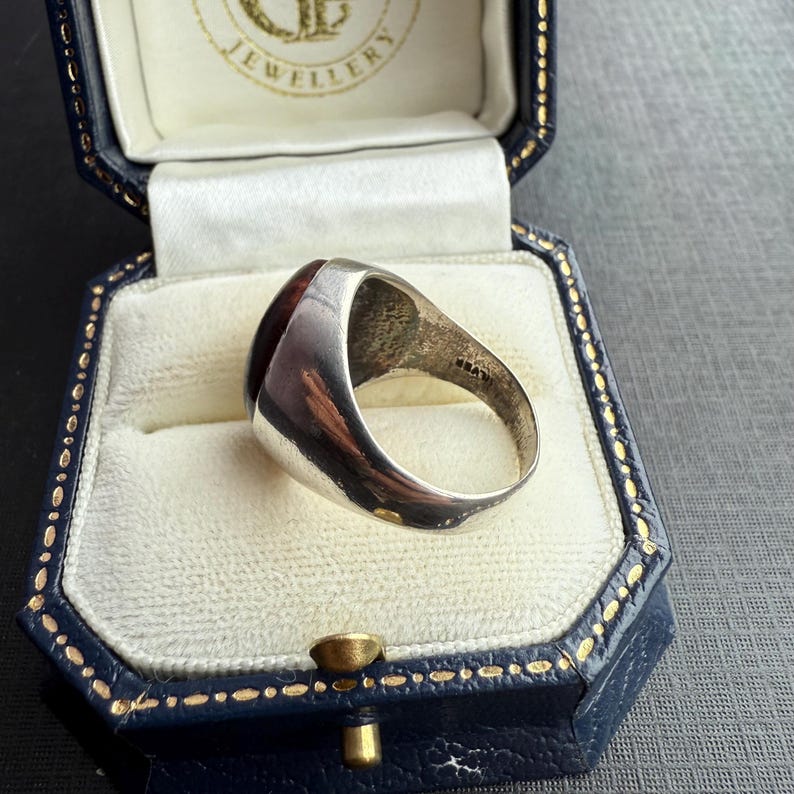May include: A silver ring featuring a large, oval, dark red gemstone. The ring is presented in a blue jewellery box with gold detailing. The ring has a polished finish and a classic design, suitable for everyday wear or special occasions.