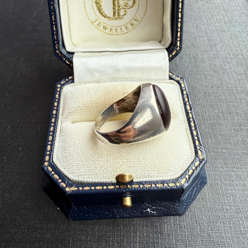 May include: A silver signet ring with a rectangular, dark red stone is presented in a blue jewellery box with a white interior. The ring is the focus, highlighting its polished metal and the rich colour of the stone.