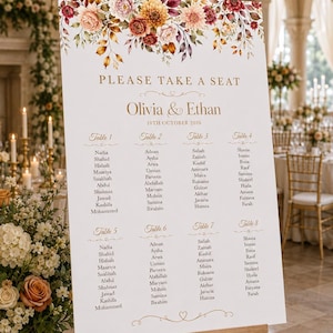 May include: A white wedding seating chart sign on a wooden easel featuring a floral arrangement of burgundy, orange, and cream flowers at the top. The text reads "PLEASE TAKE A SEAT", "Olivia & Ethan", "10TH OCTOBER 2026", and lists eight tables.