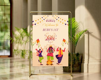 May include: A welcome sign for a first birthday party with the text "Welcome to Ruby's 1st Lohri". The sign features a colourful illustration of people celebrating Lohri with kites, a bonfire, and traditional clothing.