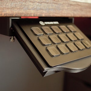 May include: A black keyboard with a silver faceplate and 16 buttons. The keyboard is mounted under a wooden desk.