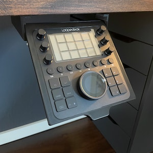 May include: A black Loopdeck controller with a circular knob, buttons, and a grid of 16 squares. The controller is mounted under a wooden desk.