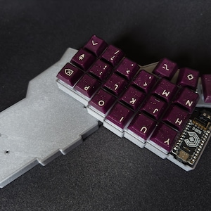 Peut inclure: Gros plan sur un petit clavier personnalisé avec des touches violettes et des lettres blanches. Le clavier est monté sur une base argentée imprimée en 3D. Le clavier est doté d'une carte de microcontrôleur fixée à l'arrière.