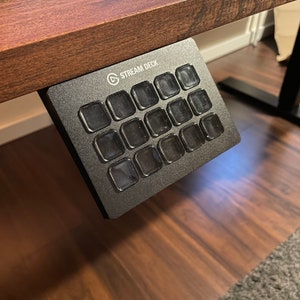 May include: A black Stream Deck with 15 round buttons, each with a clear plastic cover. The Stream Deck is mounted under a wooden desk.