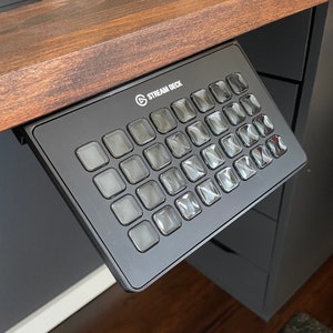 May include: A black Stream Deck with a grid of 32 customisable buttons. The buttons are made of a clear plastic material and have a slightly raised surface. The Stream Deck is mounted on a desk.