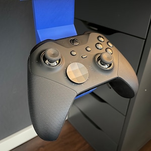 Op de afbeelding: Een zwarte Xbox-controller met een blauwe 3D-geprinte houder bevestigd aan een bureau. De controller hangt aan de houder.