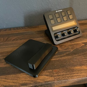 May include: A black Stream Deck mini controller with a small screen and eight buttons. The controller is sitting on a wooden surface next to a black square-shaped object.