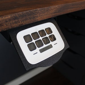May include: A white control panel with eight square buttons and a small rectangular section, mounted under a dark wooden desk. The panel is set in a black foam holder.