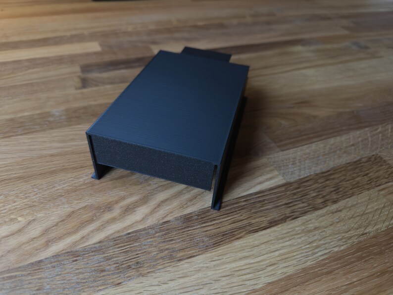 May include: A dark gray plastic box with a rectangular shape and a slightly raised lid. The box is sitting on a wooden surface.