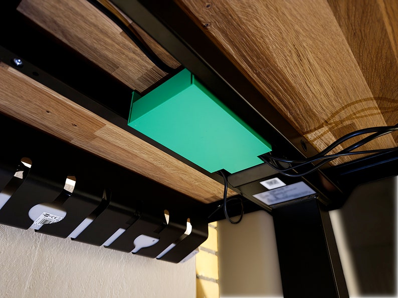 May include: A green cable management box hangs under a wooden desk. Black cords are plugged into the box and run down the side of the desk. A black cable organizer is mounted to the wall below the desk.
