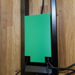 May include: A green plastic cover for cords and wires mounted on a black metal frame. The cover has a cutout at the bottom to allow cords to pass through.