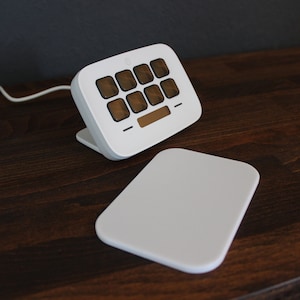 Op de afbeelding: Een wit draadloos toetsenbord met een klein display en 12 knoppen. Het toetsenbord staat op een houten oppervlak met een witte oplaadmat ernaast.