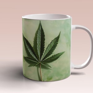 Puede incluir: Taza de cerámica blanca con un diseño de hoja de marihuana verde sobre un fondo verde y blanco.