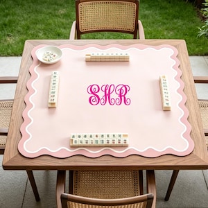 May include: A wooden mahjong table with a pink playing surface and a scalloped edge. The surface features the initials "SHR" in a cursive font. Mahjong tiles and a small white bowl are arranged on the table, surrounded by four chairs.