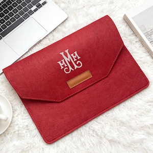 Puede incluir: Una funda roja de fieltro para portátil con un monograma blanco. La funda tiene un parche de cuero y un cierre de solapa. En la imagen también hay un portátil, una taza de café y un libro.
