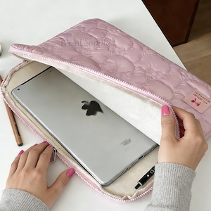 Könnte beinhalten: Eine hellrosa gesteppte Laptoptasche mit Reißverschluss, die ein silbernes Tablet enthält. Die Tasche hat ein weiches, gepolstertes Innenfutter und ein kleines Etikett mit Text und einer Schleifengrafik. Ein Bleistift und Klebeband sind sichtbar.