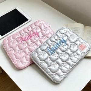 Puede incluir: Dos fundas acolchadas para tabletas, una rosa con "Paisley" en escritura rosa y otra plateada con "Lainey" en escritura azul. Ambas tienen un patrón floral. También se ven una tableta y un libro abierto sobre una superficie blanca.