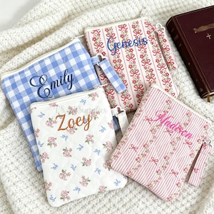 Könnte beinhalten: Vier personalisierte Reißverschlusstaschen mit gestickten Namen. Eine ist blau-weiß kariert, eine ist weiß mit rosa Blumen und zwei sind rosa gestreift mit Blumen- und Schleifenmustern. Ein weinrotes Buch mit einem Kreuz ist ebenfalls sichtbar.