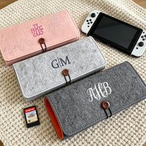 Może przedstawiać: Kolekcja filcowych etui w kolorach różowym, szarym i ciemnoszarym, każde z zapięciem na guzik i spersonalizowanym haftem monogramu. Konsola Nintendo Switch i kartridż do gier również obecne, sugerując motyw akcesoriów do gier.