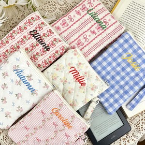 Peut inclure: Plusieurs pochettes matelassées personnalisées avec des noms brodés. Les motifs comprennent des fleurs, des rayures et du vichy. Les couleurs varient, avec du rose, du bleu et du blanc. Une pochette est à carreaux bleus et blancs.