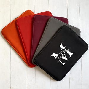 以下が含まれることがあります： オレンジ、赤、バーガンディ、グレー、黒の様々な色のノートパソコンスリーブのコレクション。黒いスリーブには、名前「michael」の白いモノグラムが付いています。各スリーブにはジッパーが付いています。