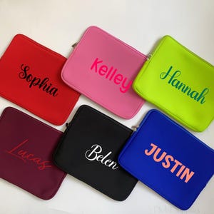 Pode incluir: Uma coleção de capas para laptop coloridas em vermelho, rosa, verde, azul, bordô e preto. Cada capa apresenta um nome em uma cor contrastante: Sophia, Kelley, Hannah, Justin, Lucas e Belen. As capas têm fecho de zíper.