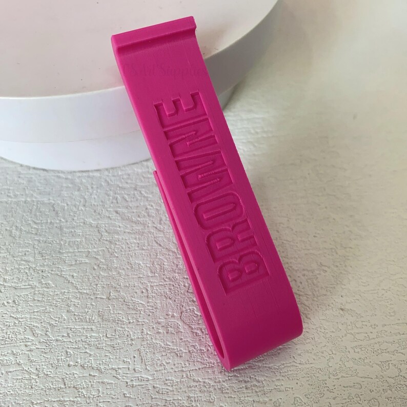 K&ouml;nnte beinhalten: Ein leuchtend pinkfarbenes Armband mit dem vertikal gepr&auml;gten Wort "BROWNE". Das Armband hat eine rechteckige Form mit einer kleinen Lasche oben und einer gebogenen &Ouml;ffnung unten. Es besteht aus einem soliden, glatten Material.