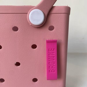 K&ouml;nnte beinhalten: Nahaufnahme einer rosa Tragetasche mit Griff und einem rosa Anh&auml;nger mit der Aufschrift "BROWNE". Die Tasche hat mehrere runde L&ouml;cher und einen wei&szlig;en Knopf am Griff.