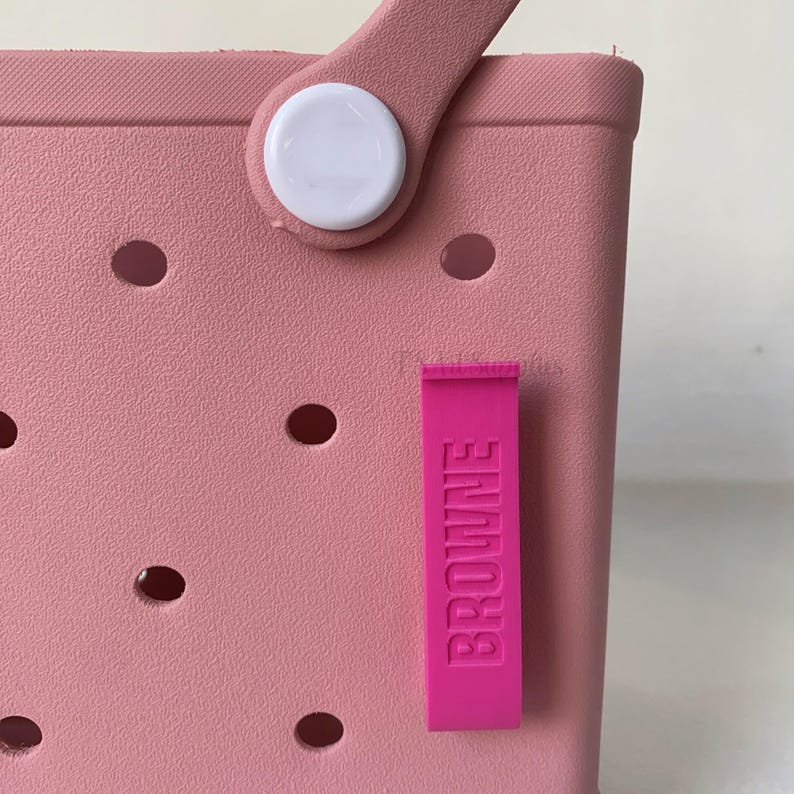 K&ouml;nnte beinhalten: Nahaufnahme einer rosa Tragetasche mit Griff und einem rosa Anh&auml;nger mit der Aufschrift "BROWNE". Die Tasche hat mehrere runde L&ouml;cher und einen wei&szlig;en Knopf am Griff.