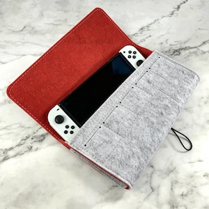 May include: A red and grey felt case with a white game console inside. The case is open, revealing the console's screen and controllers. The case has a stitched design and a black elastic closure.