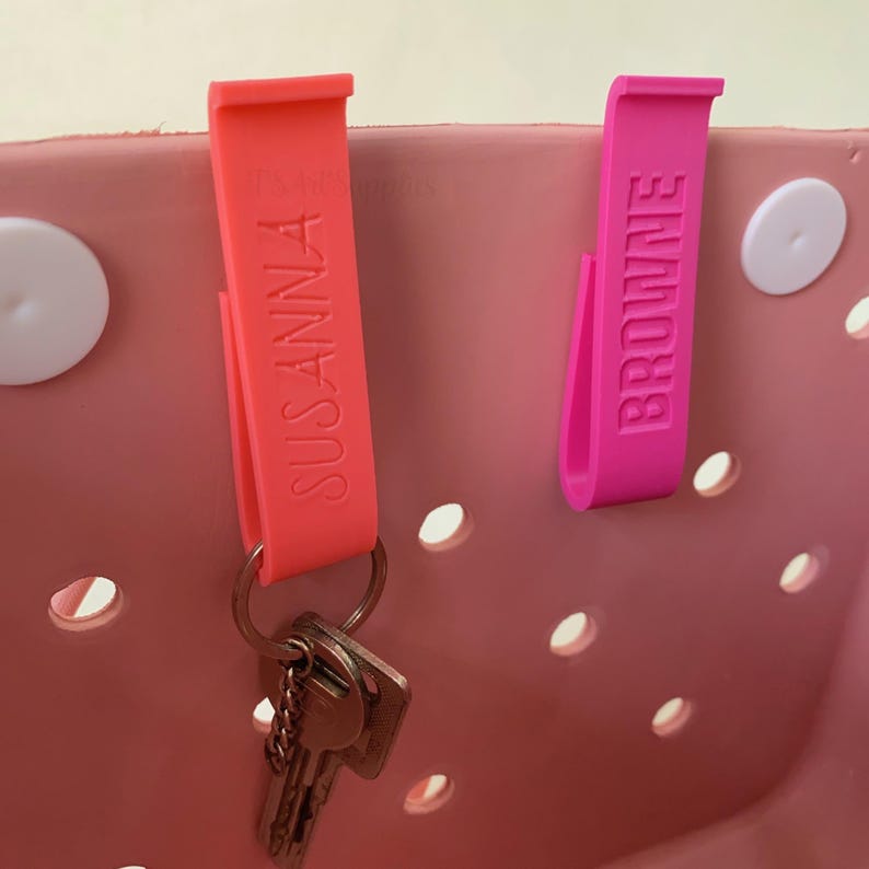 K&ouml;nnte beinhalten: Zwei personalisierte Taschenclips, einer korallenfarben und einer magenta, sind an einer rosa Tragetasche befestigt. Der korallenfarbene Clip tr&auml;gt die Aufschrift "SUSANNA" und h&auml;lt Schl&uuml;ssel. Der magenta Clip tr&auml;gt die Aufschrift "BROWNE". Die Tasche hat wei&szlig;e kreisf&ouml;rmige Akzente.