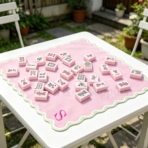 Pode incluir: Peças de Mahjong rosa com símbolos e caracteres pretos, dispostas sobre um tapete rosa com borda verde ondulada. O tapete repousa sobre uma mesa branca, com um jardim ao fundo.