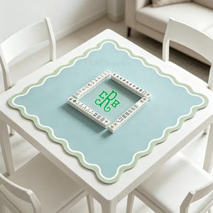 May include: A square, white table with a light blue mat featuring a scalloped edge and a green border. The mat has a green monogram and is surrounded by Mahjong tiles. The table is surrounded by white chairs.