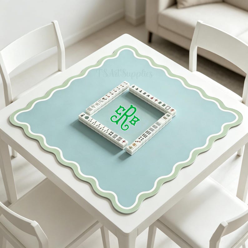 May include: A square, white table with a light blue mat featuring a scalloped edge and a green border. The mat has a green monogram and is surrounded by Mahjong tiles. The table is surrounded by white chairs.