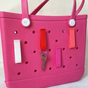 K&ouml;nnte beinhalten: Eine leuchtend rosa Tragetasche mit wei&szlig;en Griffbefestigungen und einer Reihe kreisf&ouml;rmiger Perforationen. Rote, rosa und orangefarbene Schl&uuml;sselanh&auml;nger sind an der Tasche befestigt, auf denen die Namen "Tiffany", "Browne" und "Susanna" aufgedruckt sind. Ein Schl&uuml;ssel ist am Schl&uuml;sselanh&auml;nger "Tiffany" befestigt.